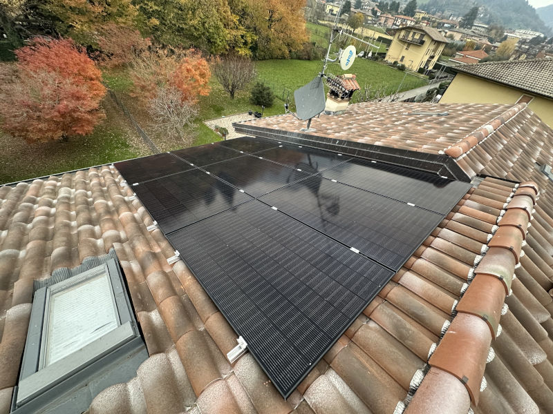 Impianto fotovoltaico con pannelli neri installati su un tetto in coppi, con lucernario e abitazioni circostanti. Impianto fotovoltaico con pannelli neri installati su un tetto in coppi, con lucernario e abitazioni circostanti.